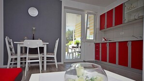 A modern kitchen with red cabinets, a dining area with white chairs, and a large window with blinds. - Apartment Ines (Vodice)