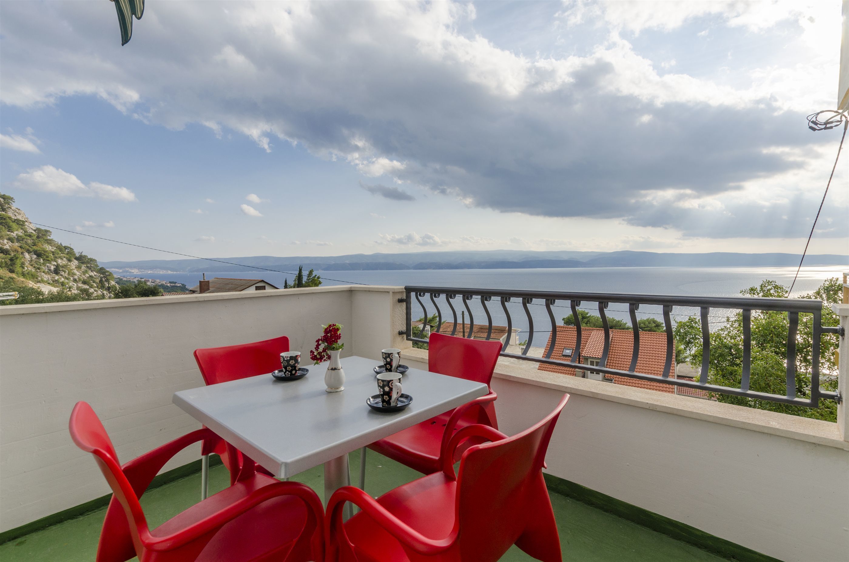 Un balcone con tavolo e sedie rosse, affacciato su uno specchio d'acqua e colline lontane.
