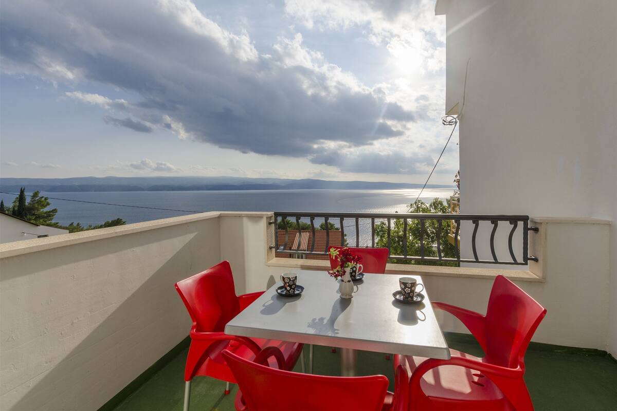 A balcony with a table and chairs, a vase with flowers, and a view of the sea and mountains.