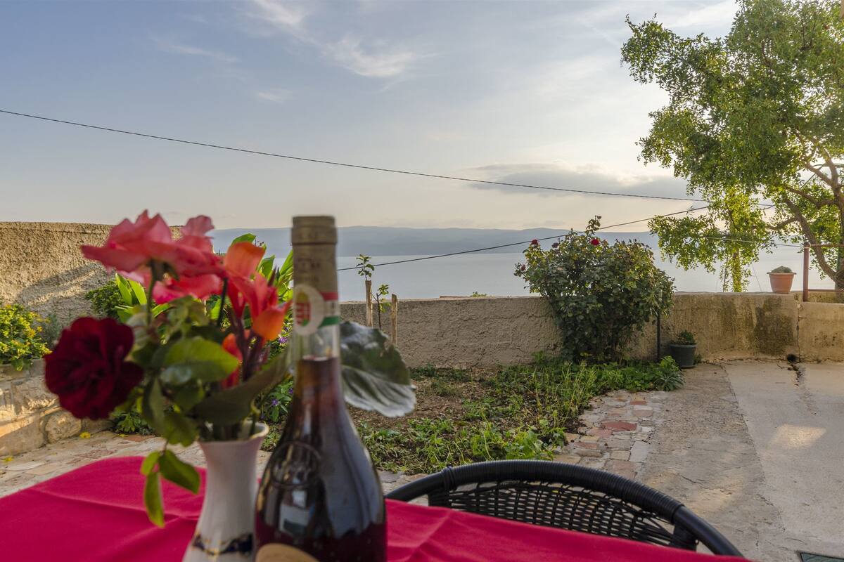 A table with a red tablecloth, a bottle, and flowers. A view of the sea and a stone wall is visible in the background.