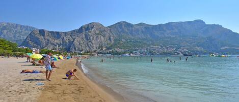 A beach with clear water, mountains in the background, and people swimming and sunbathing.