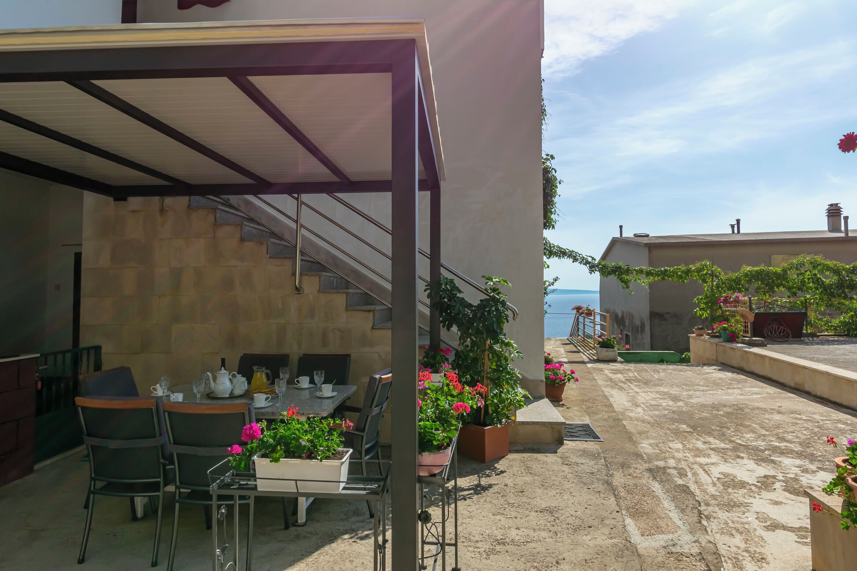 A covered outdoor seating area with tables and chairs, potted plants, and a view of the sea.