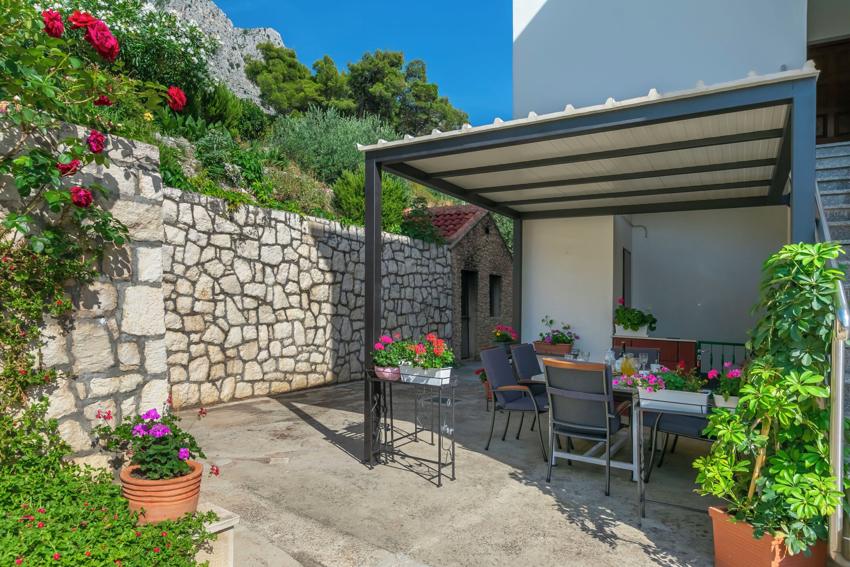 Un patio al aire libre techado con paredes de piedra, plantas en macetas y una mesa con sillas.