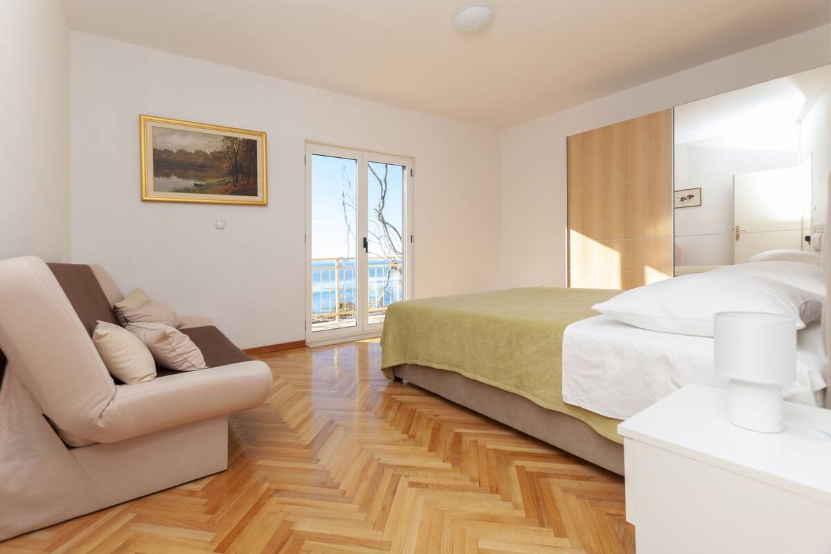 A bedroom with a bed, a sofa, a painting, and a view through a glass door.
