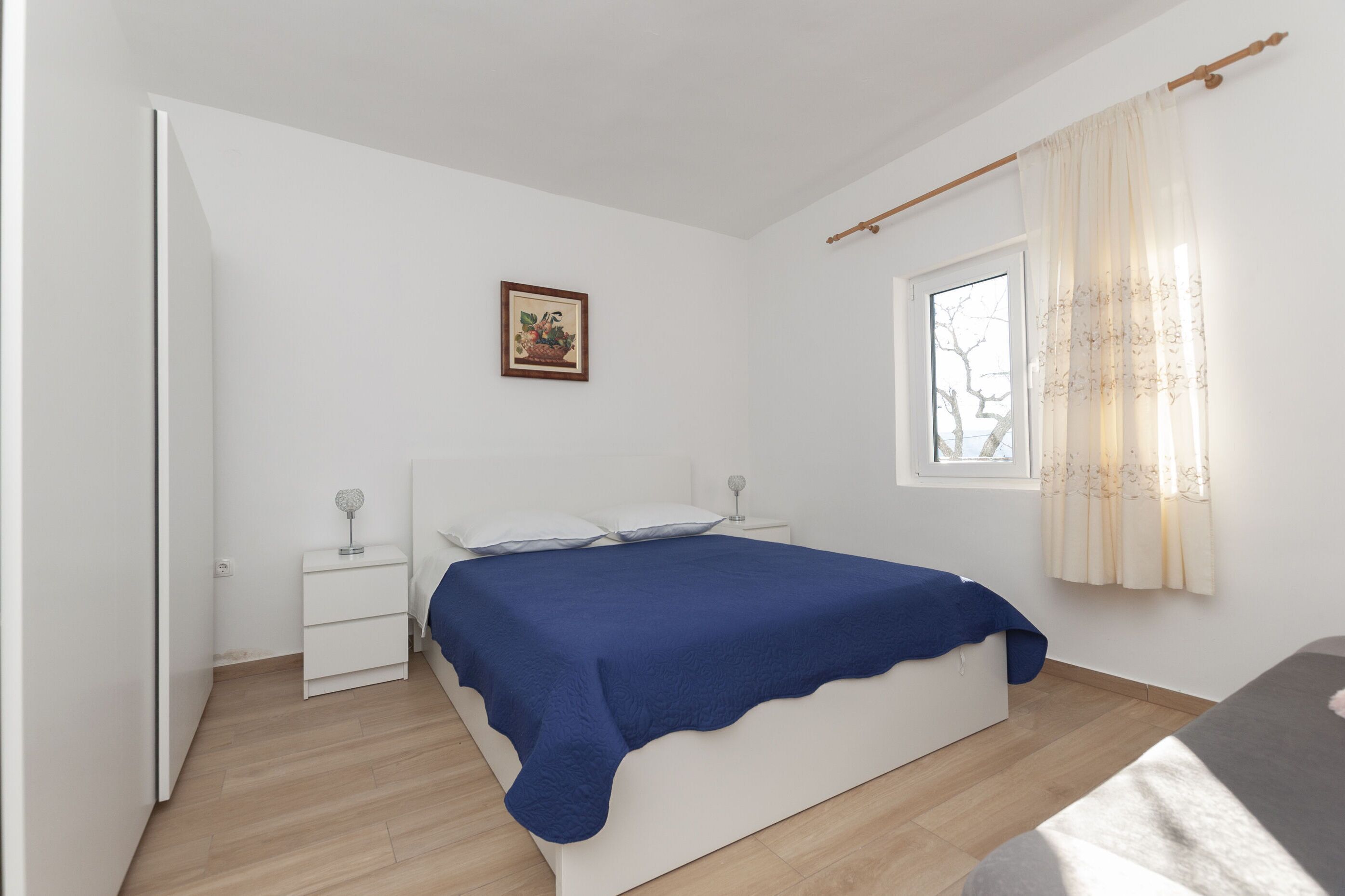 A bedroom with a bed, a bedside table, a nightstand, a window with curtains, and a wall-mounted picture.