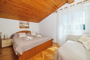 Una camera da letto con soffitto in legno, un letto con copripiumino color beige, un comodino con una lampada e una finestra con tende trasparenti.