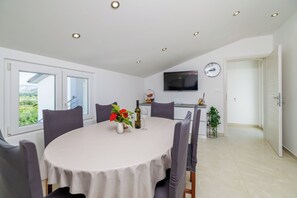 Zona pranzo con tavolo rotondo, sedie, un vaso con fiori, una bottiglia di vino e una televisione appesa al muro.