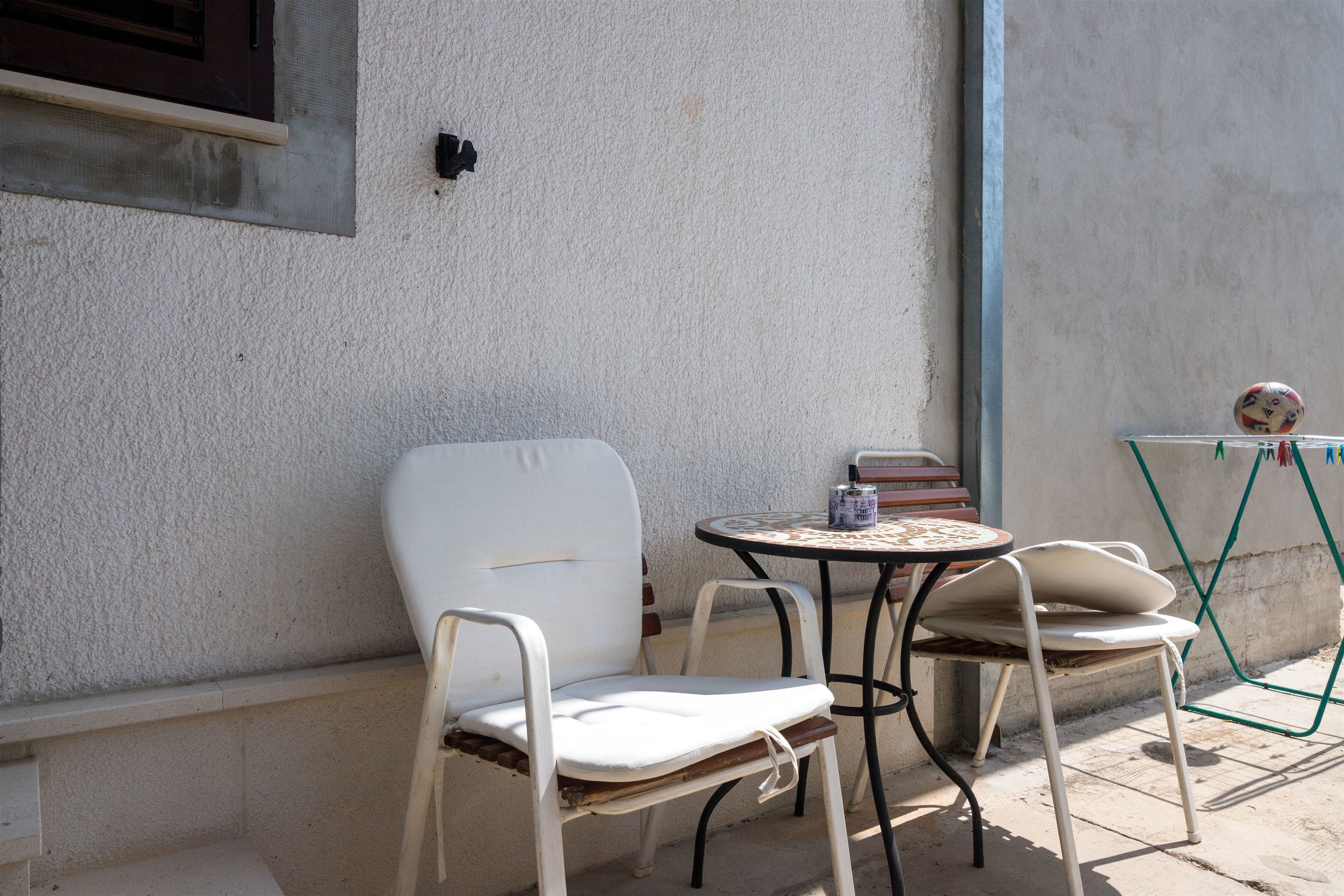 A patio area with two white metal chairs and a small round table, a green folding chair, and a wall-mounted shelf with books and a decorative item.