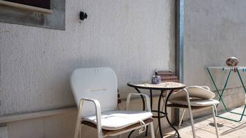En uteplats med två vita metallstolar och ett litet runt bord, en grön hopfällbar stol och en vägghylla med böcker och en dekorativ prydnad.
