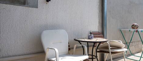 A patio area with two white metal chairs and a small round table, a green folding chair, and a wall-mounted shelf with books and a decorative item.