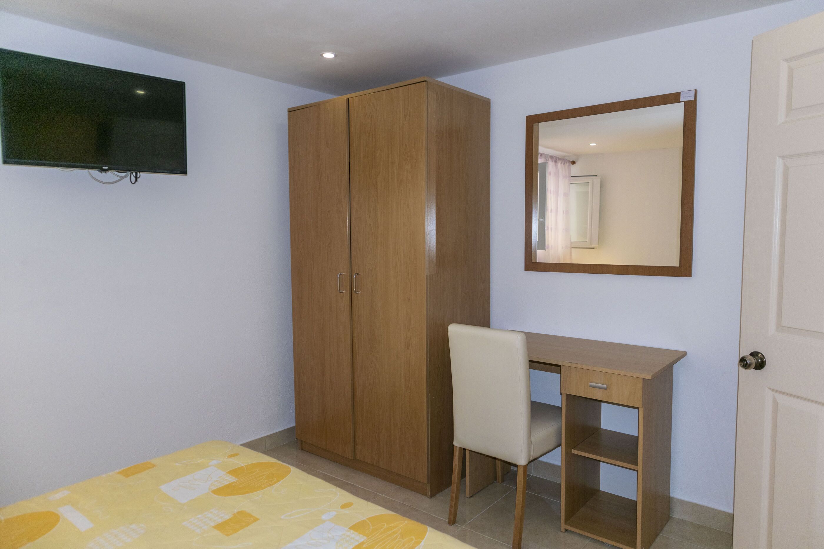 A bedroom with a bed, a wooden wardrobe, a desk with a chair, a television, and a mirror.