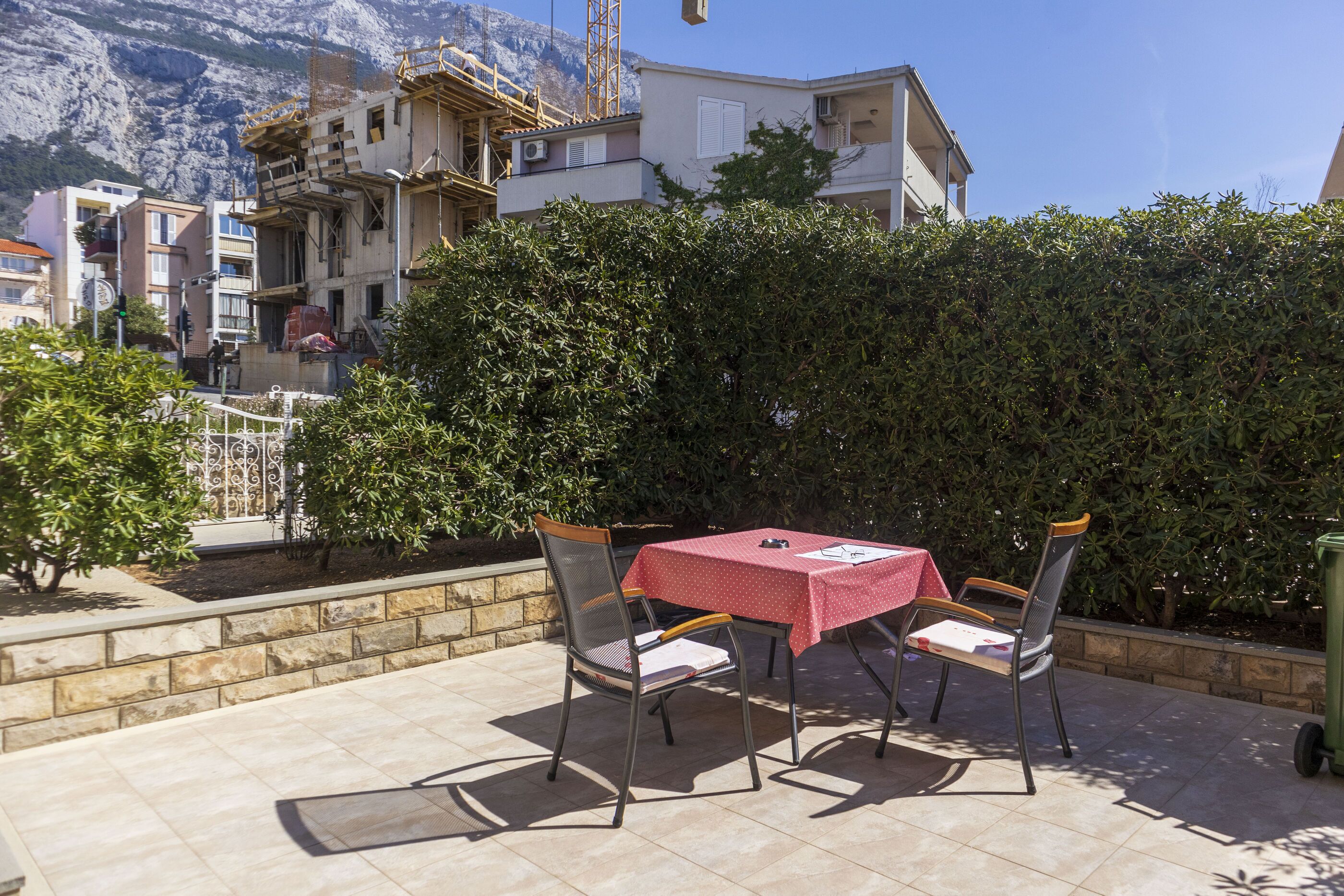 Eine Terrasse im Freien mit Tisch und zwei Stühlen, umgeben von einer Hecke und einer Steinmauer.