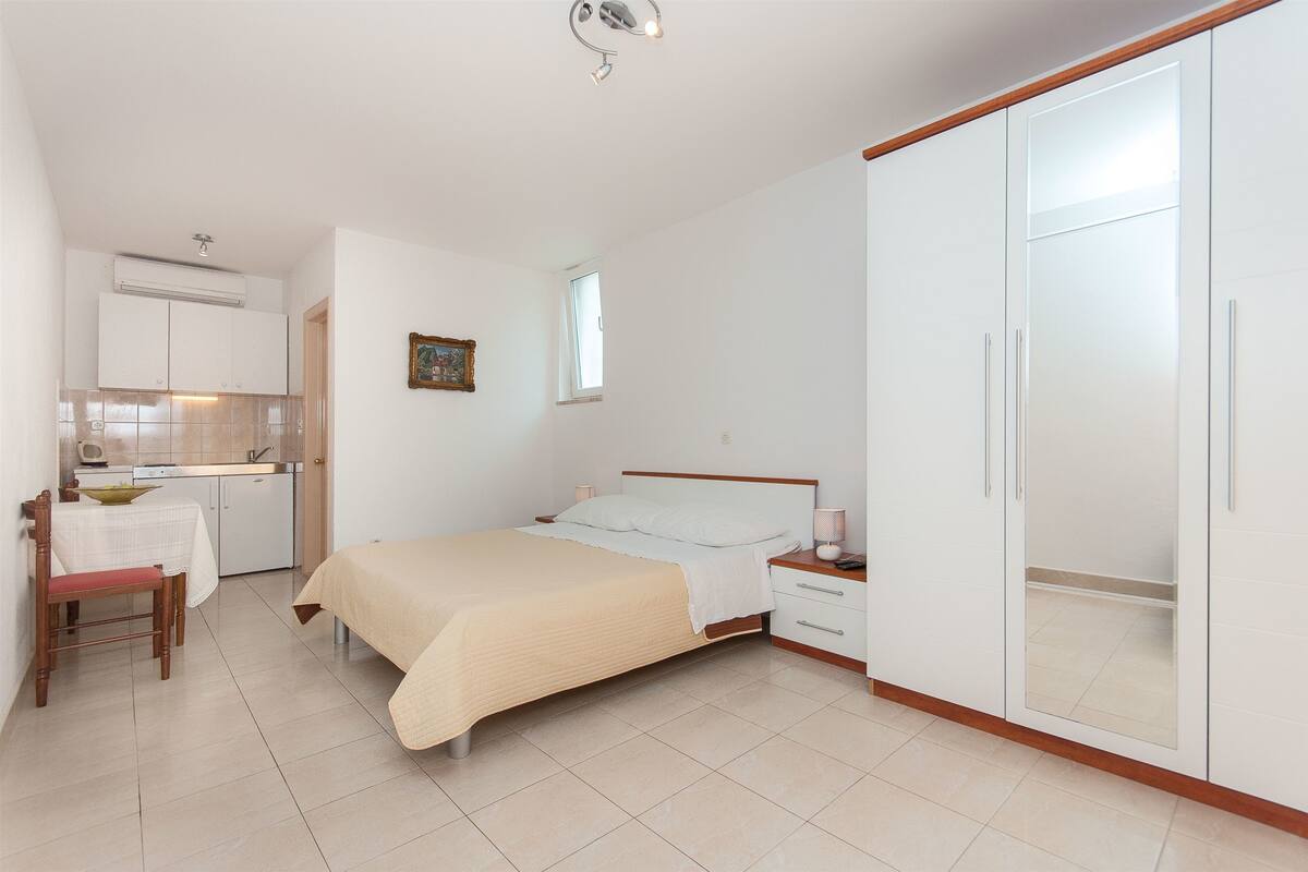 A bedroom with a bed, bedside table, and wardrobe. There is a small kitchenette with a table and chairs.