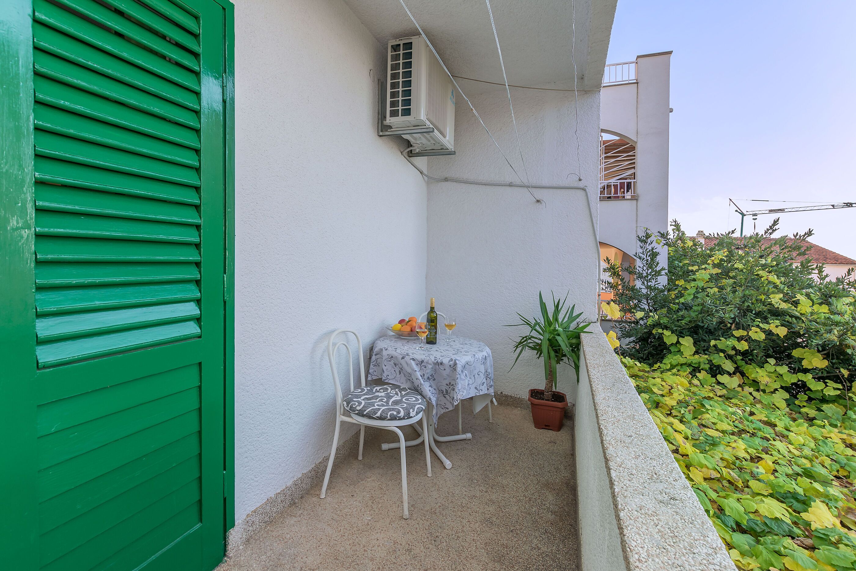 A balcony with a green shutter, a white table with a patterned tablecloth, two chairs, a potted plant, and a small table with a bottle and glasses.