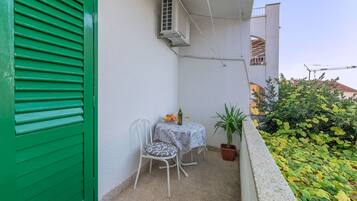 A balcony with a green shutter, a white table with a patterned tablecloth, two chairs, a potted plant, and a small table with a bottle and glasses.