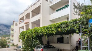 A multi-story building with a covered parking area, a staircase leading to the entrance, and a view of the surrounding area. - Apartments and Room Nevenka (Makarska)
