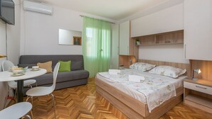 A modern hotel room with a wooden floor, a bed with floral bedding, a grey sofa, a dining table with chairs, and a TV mounted on the wall. - Apartments and Room Nevenka (Makarska)