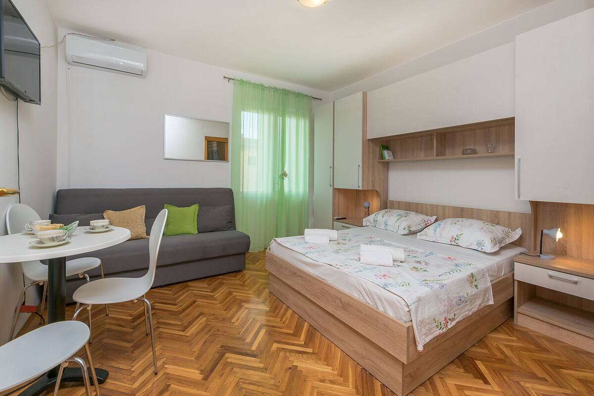 Ein modernes Hotelzimmer mit Holzboden, einem Bett mit floralem Bettzeug, einem grauen Sofa, einem Esstisch mit Stühlen und einem an der Wand montierten Fernseher.