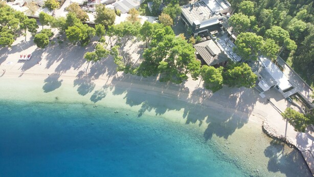 An aerial view of a coastal area with clear blue waters, a sandy beach, and a cluster of buildings with trees nearby. - Apartments and Room Nevenka (Makarska)