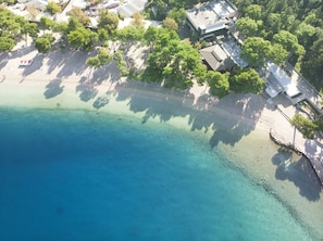 An aerial view of a coastal area with clear blue waters, a sandy beach, and a cluster of buildings with trees nearby.