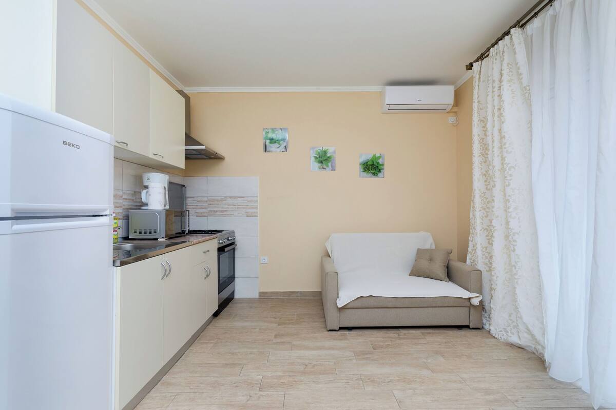 A compact kitchen with a refrigerator, microwave, and stove. There is a beige sofa and a white curtain in the living area.