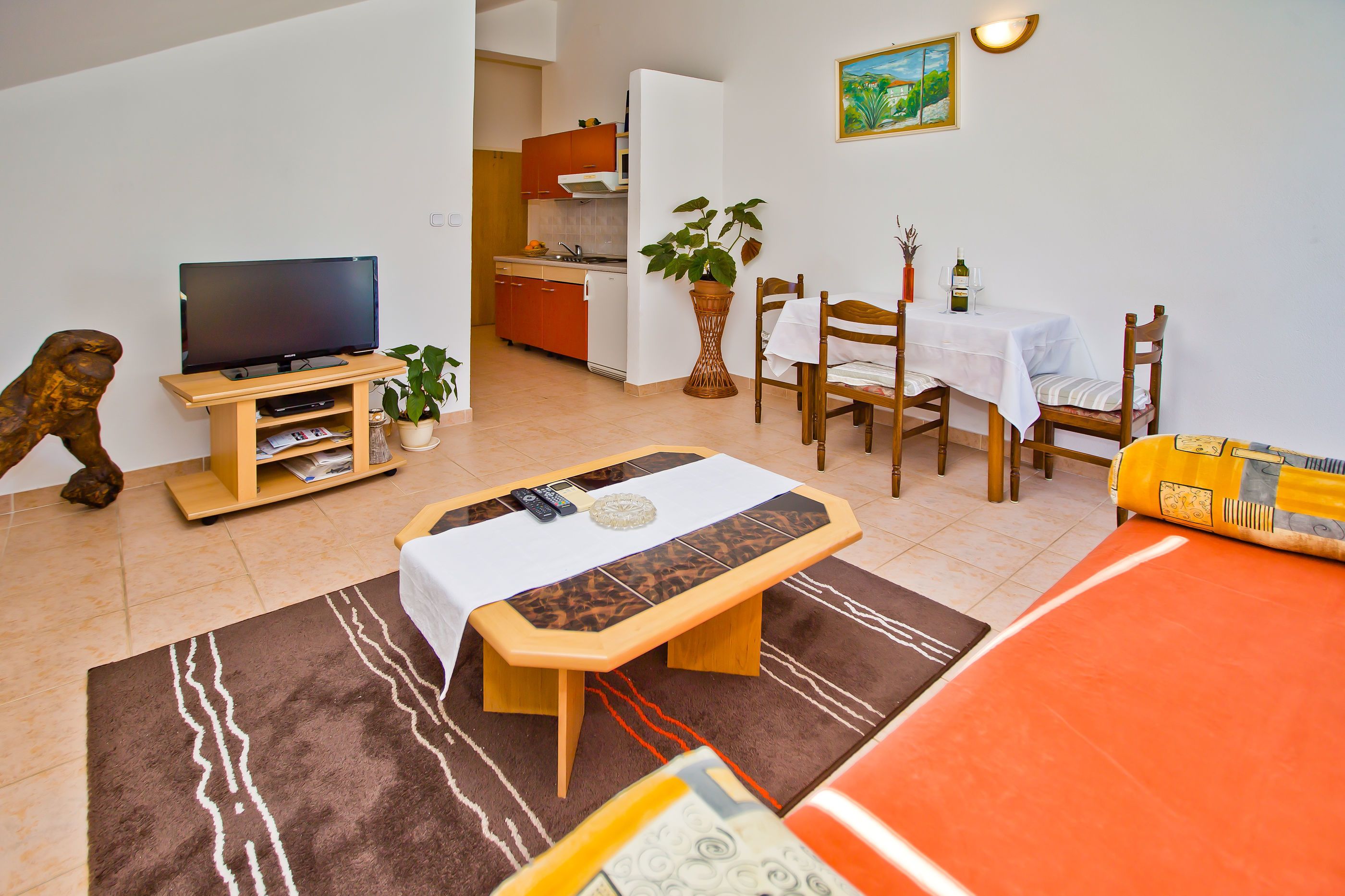 A living room with a TV, a dining table, chairs, a bench, and a kitchen in the background.