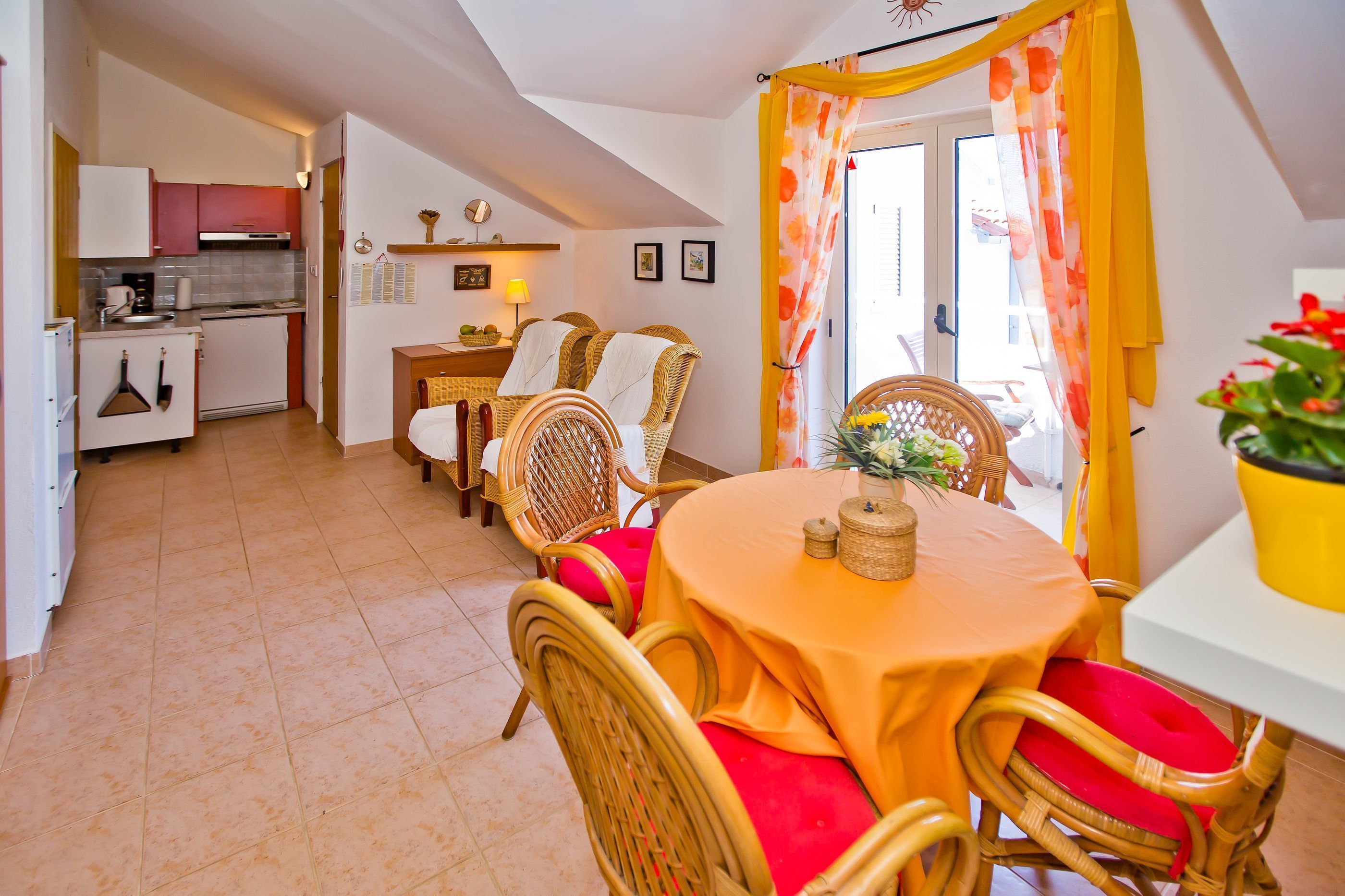 Un espace de vie compact doté d’un coin repas avec des chaises en osier et une table recouverte d’une nappe jaune. On aperçoit à l’arrière-plan une cuisine équipée d’appareils modernes et une petite table à manger.