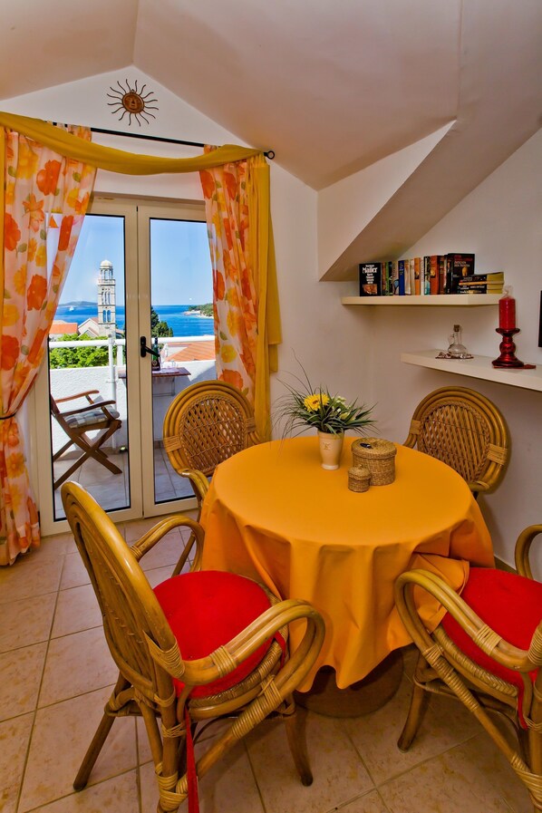 Un coin repas avec des chaises en osier, une table ronde recouverte d’une nappe jaune, une plante en pot et une vue sur la mer par une porte vitrée.