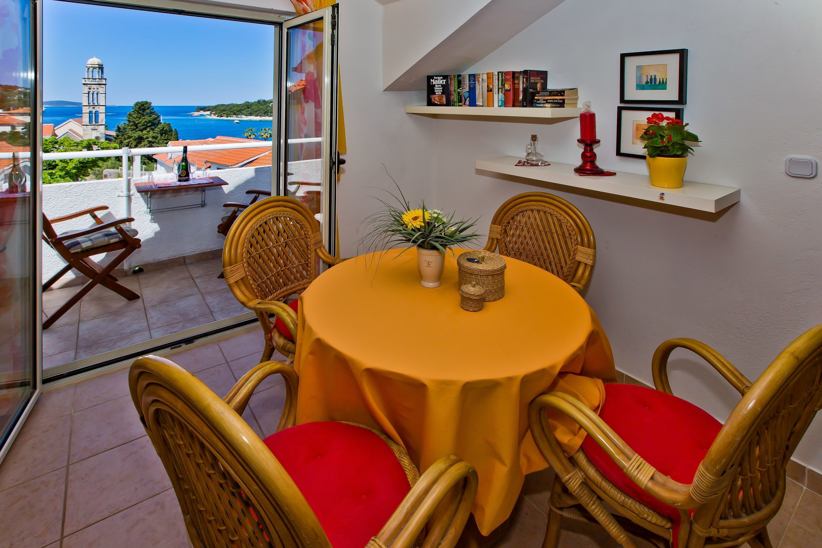 Un comedor con una mesa dispuesta para cuatro personas, rodeado de sillas de mimbre con cojines rojos, un mantel amarillo y vistas al mar y a una torre a través de un ventanal.