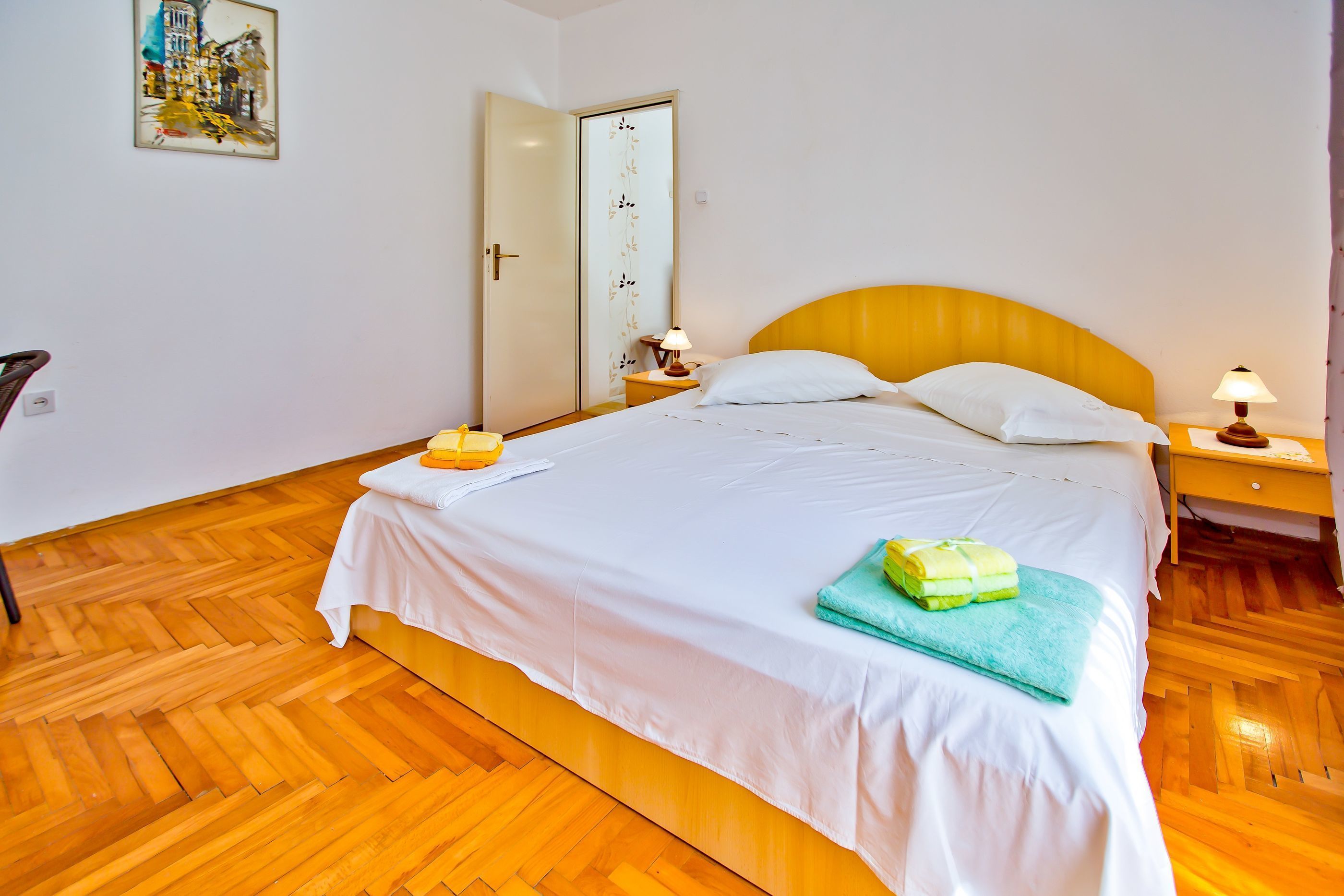 A hotel room with a wooden herringbone floor, a bed with white bedding, two bedside lamps, and a door with a floral design.