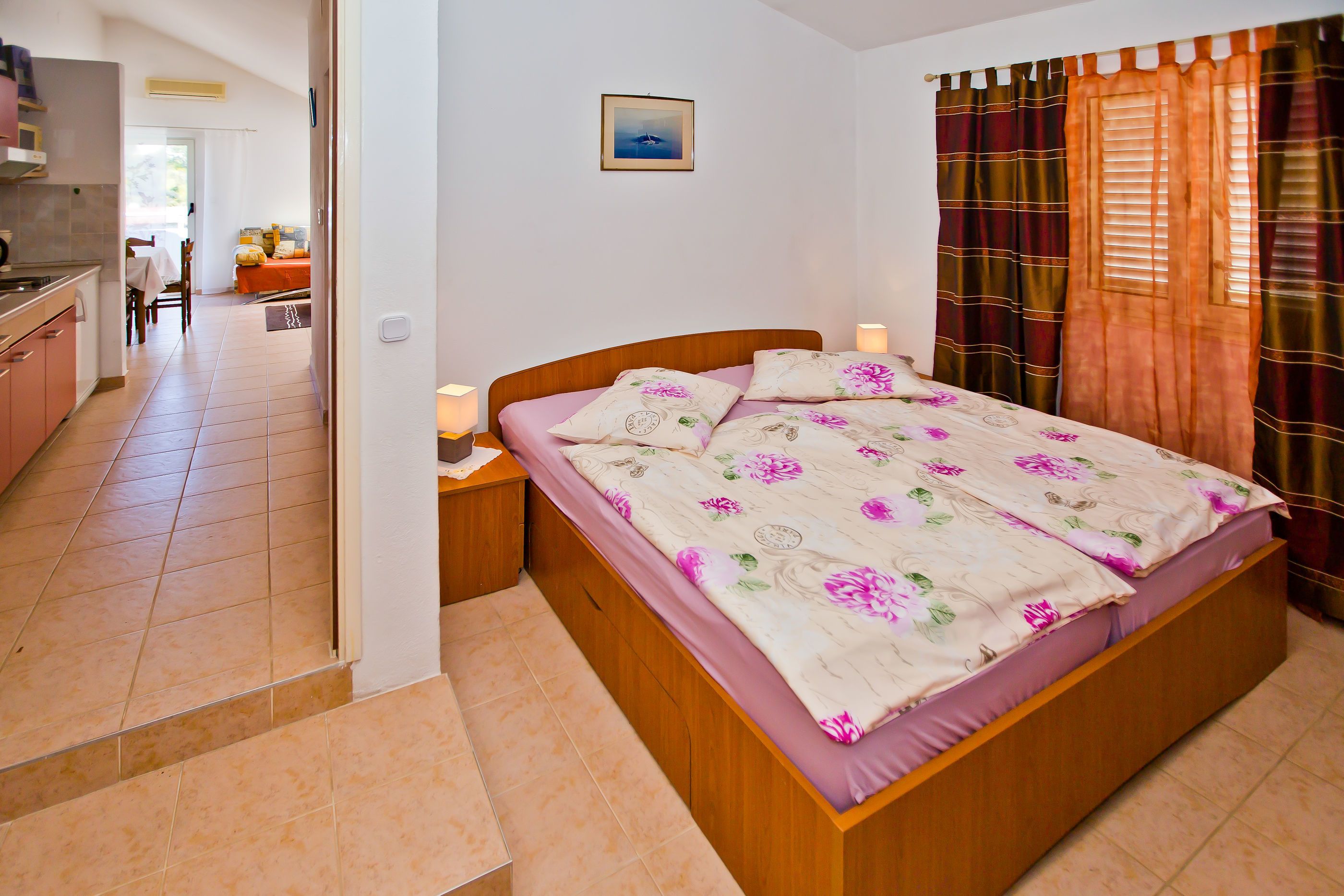 A bedroom with a wooden bed, a small bedside table, a nightstand, a window with curtains, and a view of a kitchen and dining area.