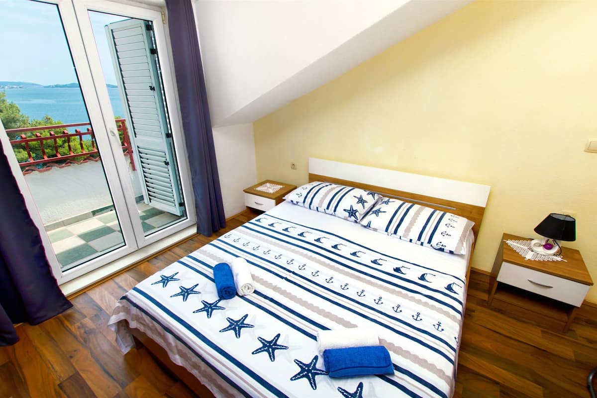 A bedroom with a bed, a nightstand, a lamp, and a view of the sea through a sliding door.
