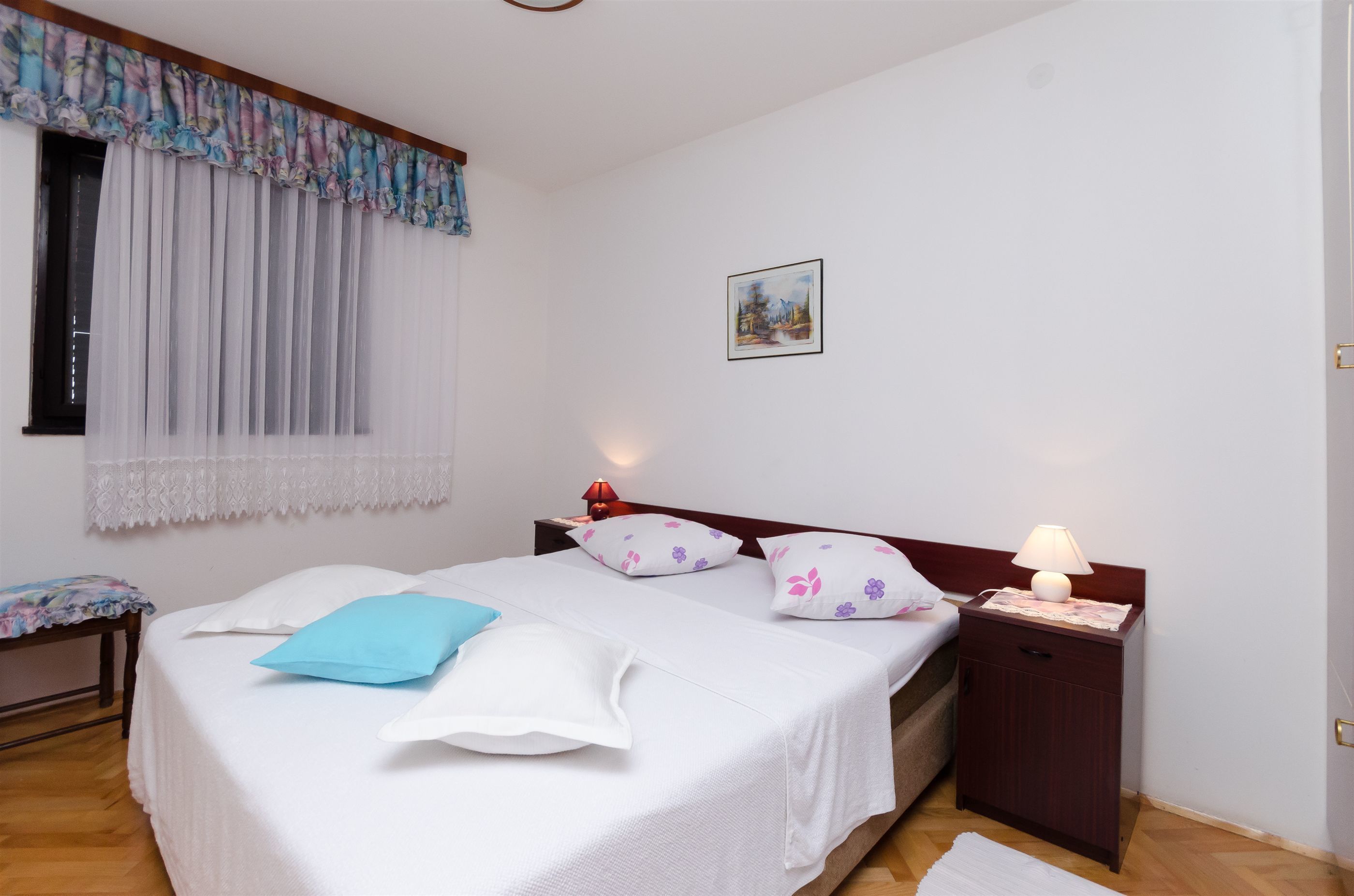 A neatly made bed with a white and blue pillow, a bedside table with a lamp, and a window with sheer curtains.