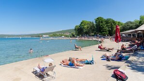 A beachside setting with sun loungers, umbrellas, and people relaxing by the water. - Apartments Branka (Punat)