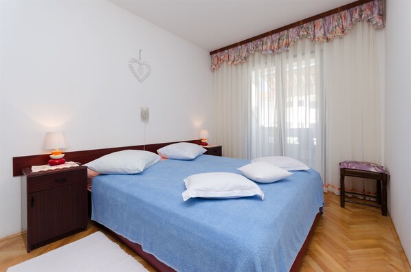 Un lit bien fait, avec une couvre-lit bleue et des oreillers blancs, une table de chevet en bois avec une lampe, et une petite table avec un coussin.