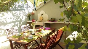 Un coin repas avec une table dressée pour quatre personnes, des chaises, une plante suspendue et une étagère fixée au mur, sur laquelle on trouve des fruits et un distributeur d’eau.