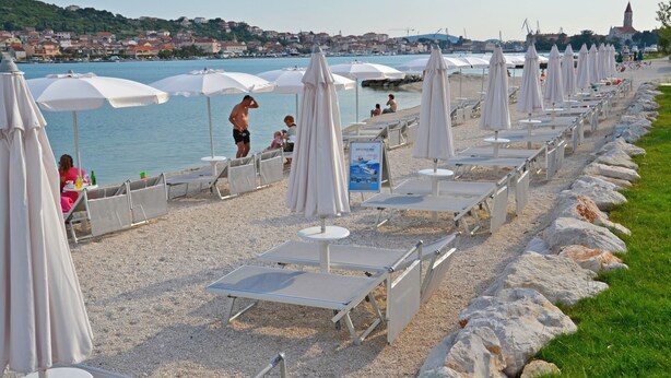 A beach with sun loungers and umbrellas, a rocky shoreline, and a view of a town across the water. - Apartment Anica (Trogir)