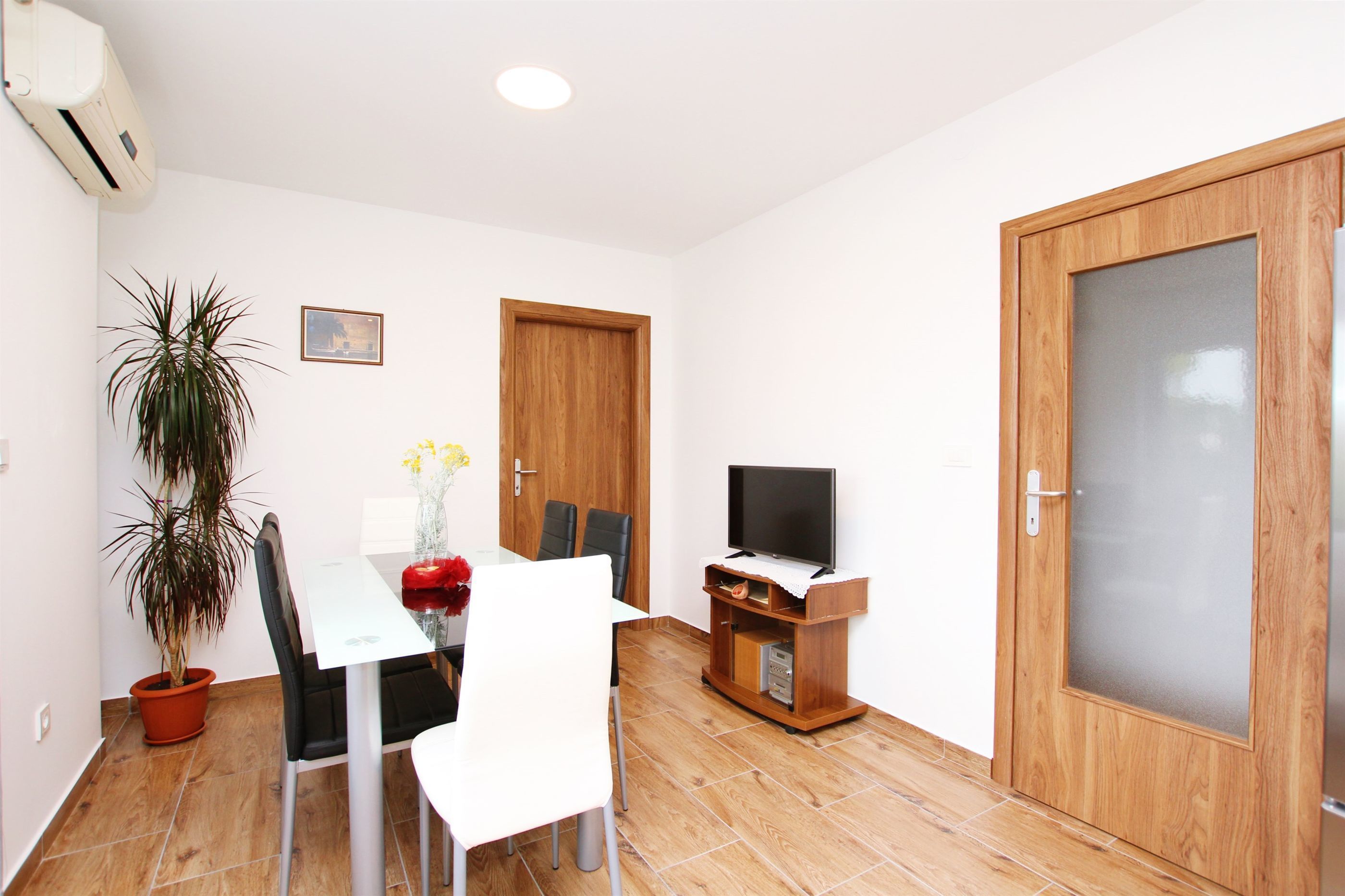 A compact living space with a dining table, chairs, a potted plant, a television on a wooden stand, and a large wooden door.