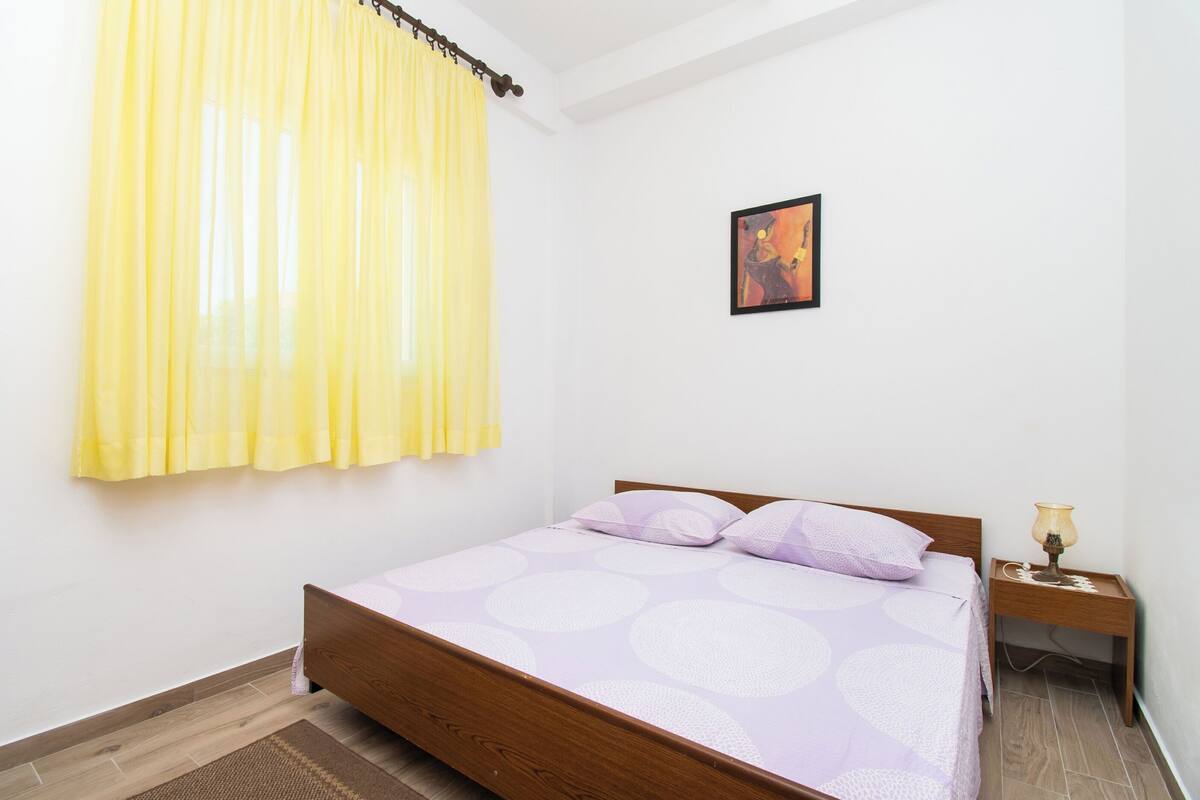 A bedroom with a wooden bed, a nightstand with a lamp, a painting on the wall, and yellow curtains.