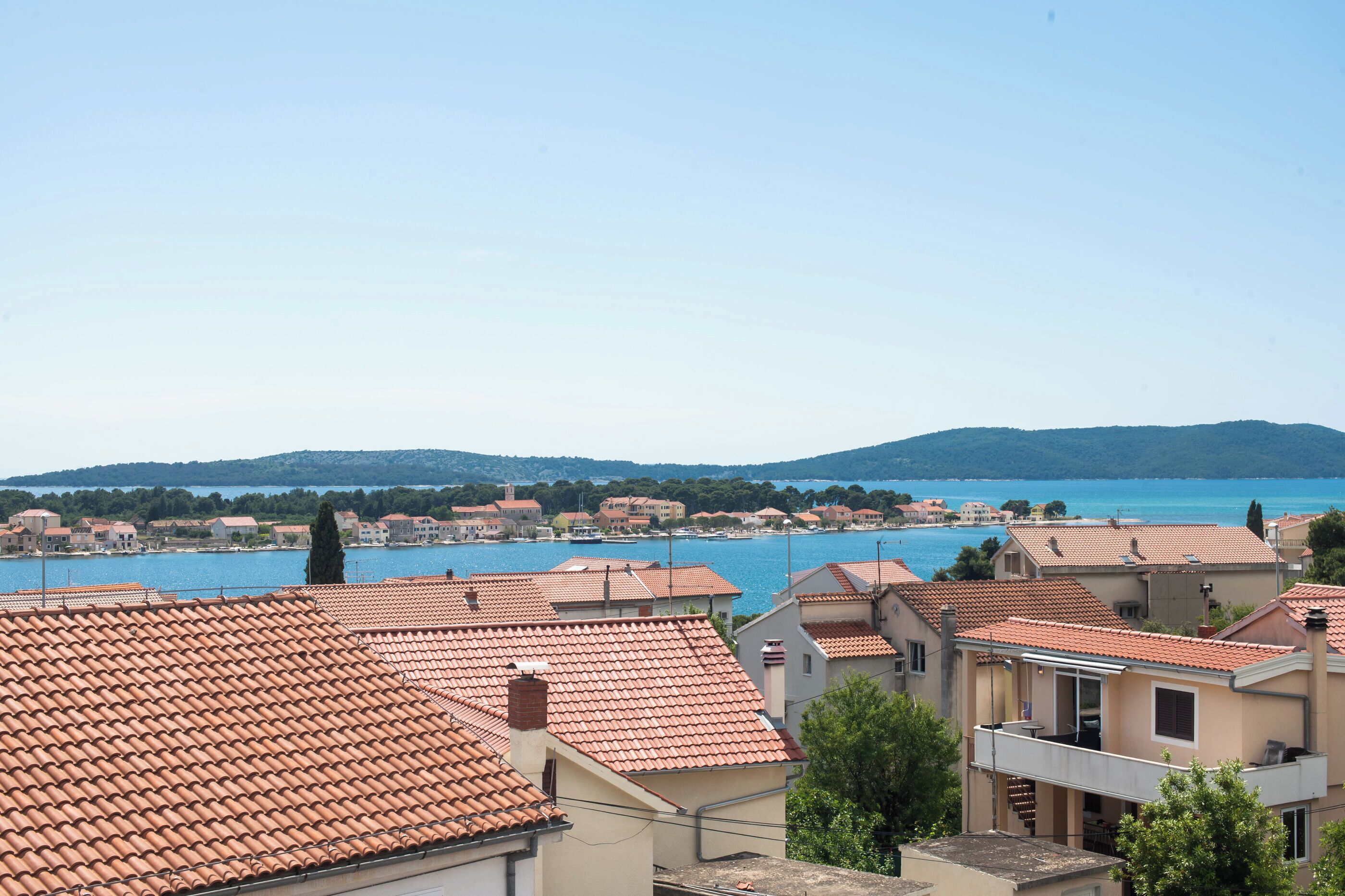 A coastal residential area with terracotta-tiled roofs, a clear blue sea, and hills in the background.