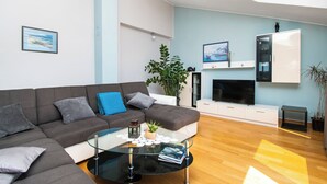 A modern living room with a grey sectional sofa, a glass coffee table, a flat-screen TV mounted on the wall, and a white cabinet with a plant on top. - Apartments Cace (Brodarica)