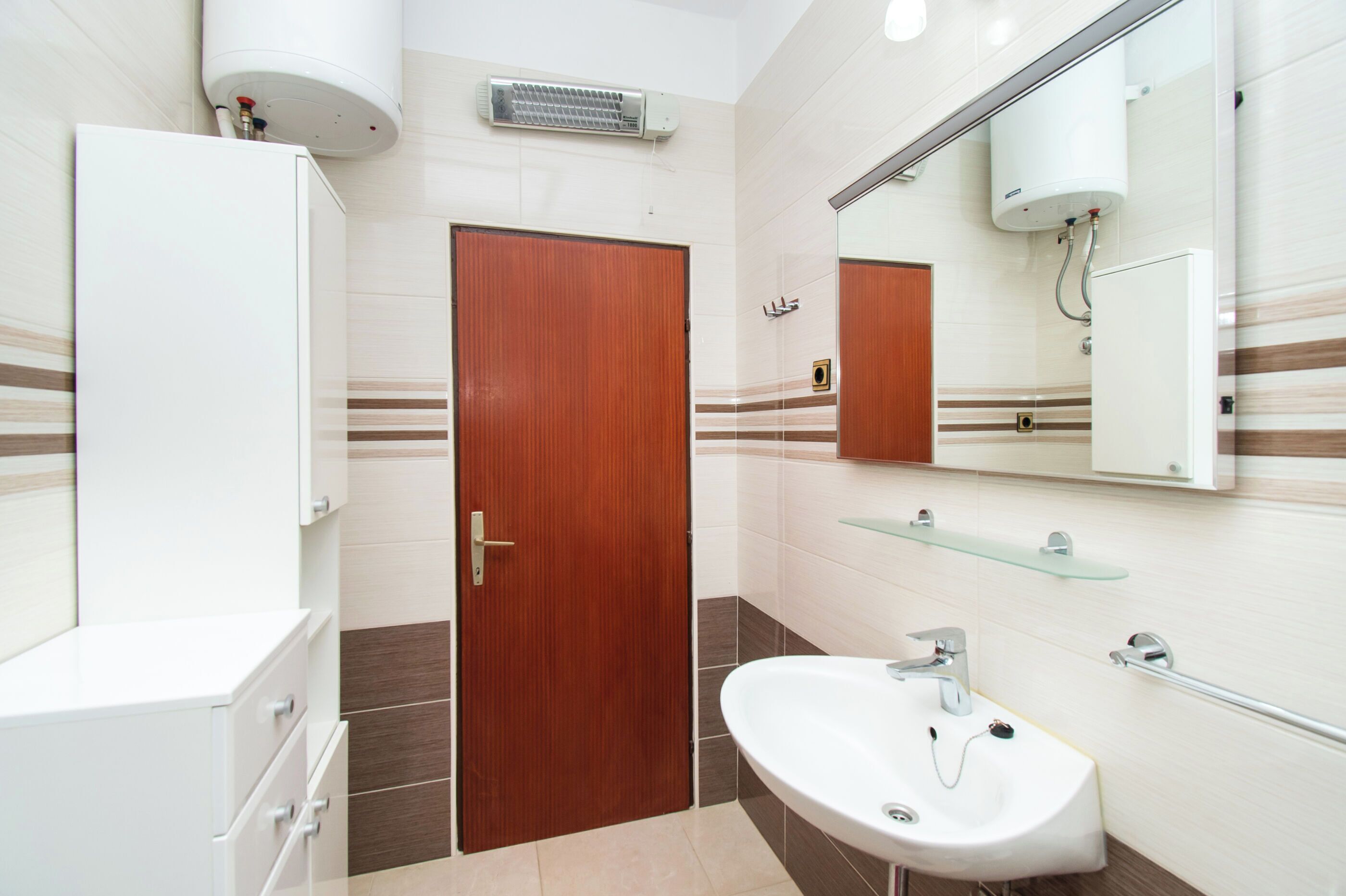 Un baño moderno con puerta de madera, lavamanos blanco, estante de vidrio y espejo.