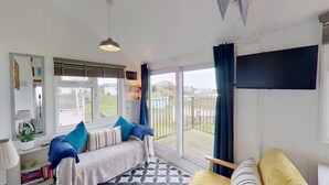 A cozy living room with a sofa, a coffee table, and a TV mounted on the wall. There is a large window with a view of the outside. - F17 Up-A-Bank, Riviere Towans (Hayle)