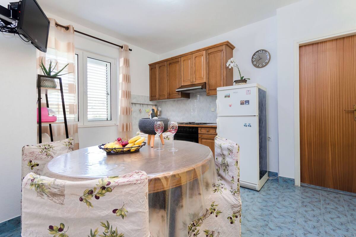 A compact kitchen with wooden cabinets, a refrigerator, and a dining table with a fruit bowl and wine glasses.