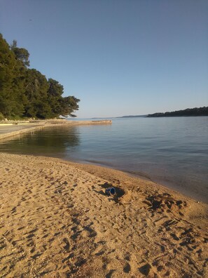 A sandy beach with a calm sea, a pier, and a forested area in the background. - Apartments Marotin (Palit)