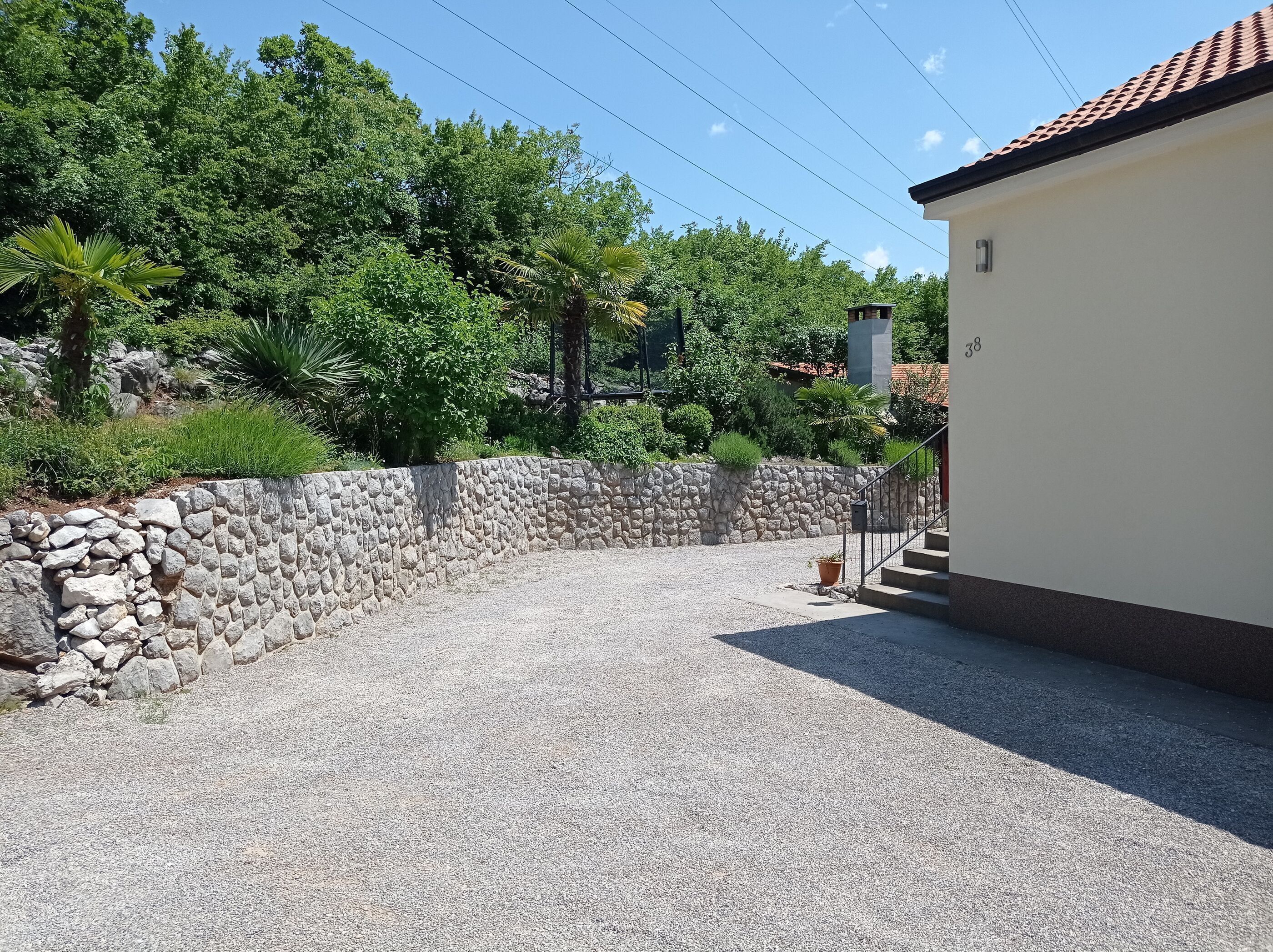 A gravel driveway leading to a building with a stone wall and a small garden area with various plants.