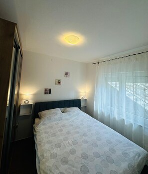 Ein Schlafzimmer mit einem Bett, zwei Nachttischlampen, einem Fenster mit transparenten Vorhängen und einer Deckenleuchte.