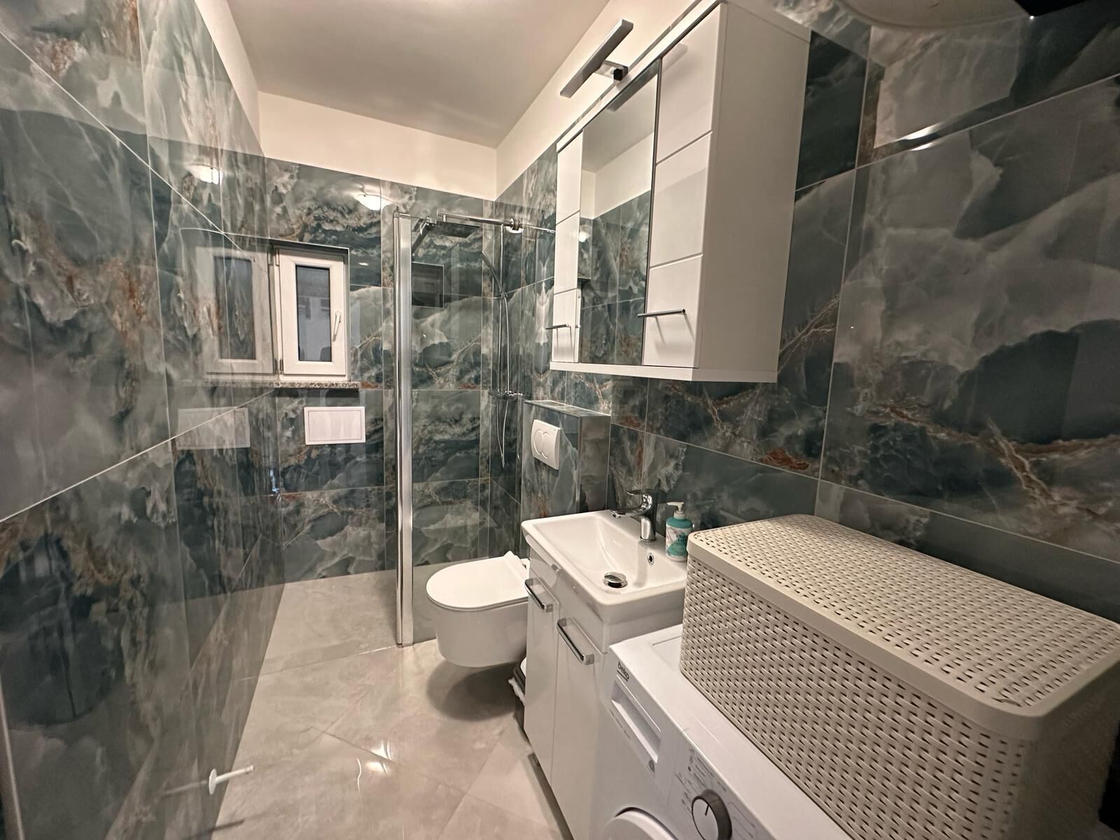A modern bathroom with a glass shower enclosure, a toilet, a sink, and a washing machine.