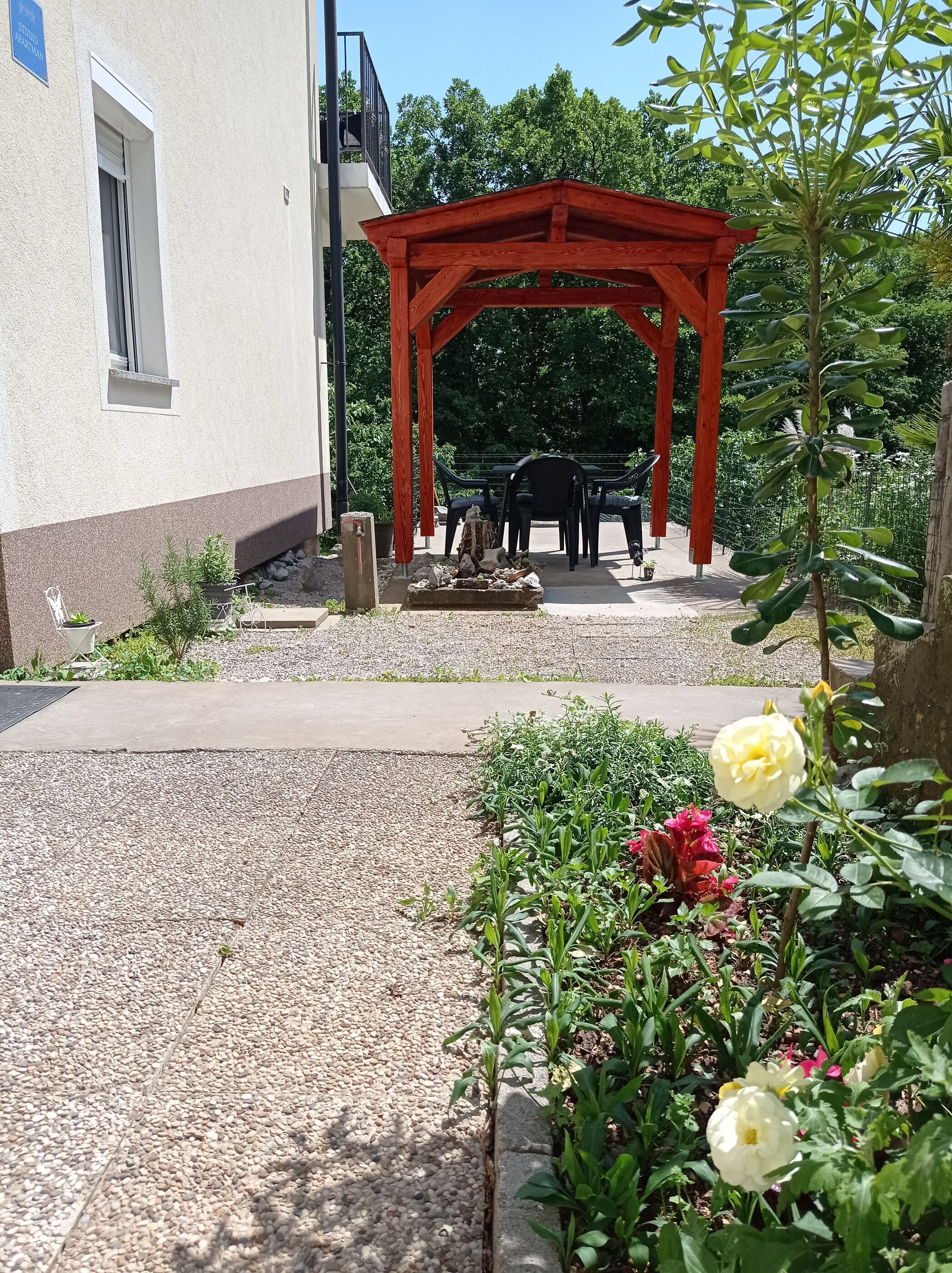 Een tuin met een houten pergola, een zithoek met stoelen en verschillende planten en bloemen.