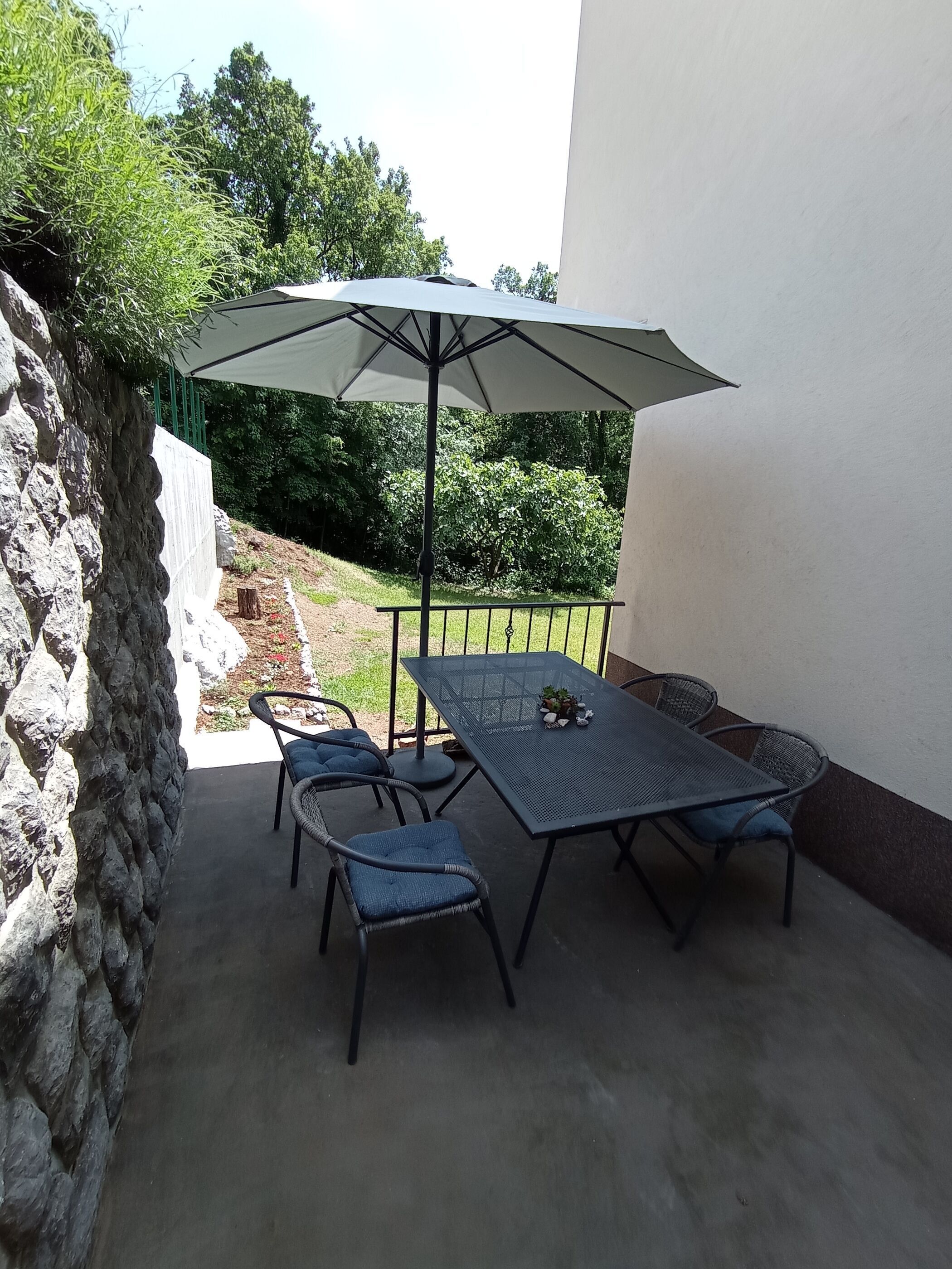 Eine Terrasse im Freien mit Tisch, Stühlen und einem großen Sonnenschirm, mit Blick auf einen Garten.